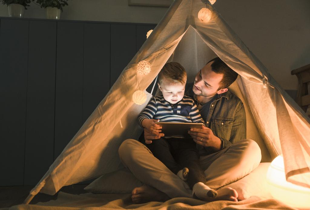 image of father and son inside a blanket fort with twinkle lights on reading a book from a tablet